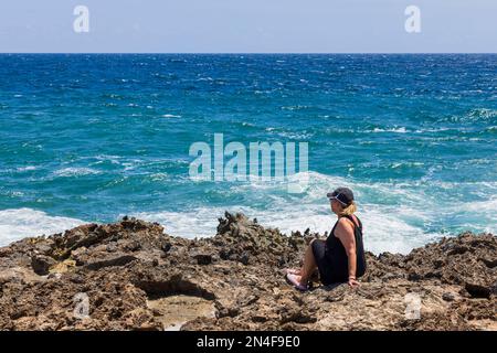 Wunderschöne Aussicht auf eine Frau, die auf Felsen an der Nordküste der Insel Aruba sitzt und die türkisfarbenen Wellen des Atlantischen Ozeans betrachtet. Stockfoto