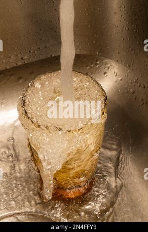 Vertikales Bild von Wasser, das über ein Trinkglas in einem Edelstahlwaschbecken fließt. Stockfoto
