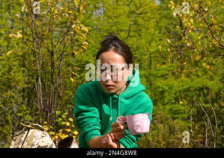 Eine aufmerksame Yakut-Asiatin mit Gläsern, die neben einem Rucksack im wilden Wald von Yakutia saß und eine Tasse Tee trank. Stockfoto