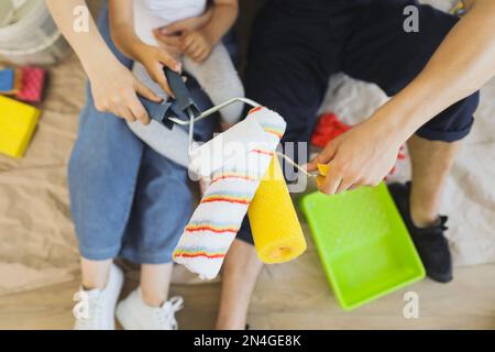 Gekürzte Nahansicht Hände von Familie, Vater, Mutter und kleiner Tochter spielen, während sie in ihrem hellen Zimmer renovieren und ihre Hände mit Malerwalzen zusammenlegen. Stockfoto