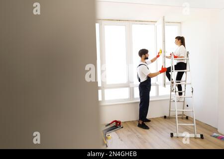 Ein wunderschönes weißes Paar, Frau und Mann, die zusammen das Fenster putzten. Arbeitende Familien verwenden Spray beim Waschen von Fenstern, arbeiten zusammen. Stockfoto