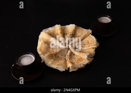 Foto-Pfannkuchen in einem Kreis auf einem Teller und zwei Tassen mit einem Getränk auf schwarzem Hintergrund Stockfoto