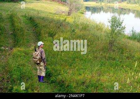 Yakut asiatischer Touristenangler in Schutzkleidung mit einem Rucksack und einer Angelrute in der Hand auf dem Pfad in der Wildnis Stockfoto