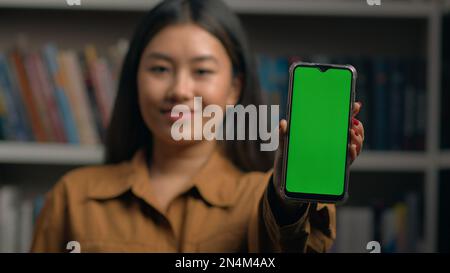 Lächelnde koreanische Geschäftsfrau mit Telefon in der Hand, die ein Smartphone mit grünem Touchscreen und Touchscreen-Display mit Chroma-Key-Werbung zeigt Stockfoto