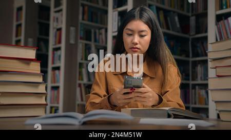 Junge, seriöse asiatische Studentin, die in der Bibliothek am Schreibtisch sitzt und Informationen im Internet mit einem Mobiltelefon sucht, um sich auf die Prüfung vorzubereiten, nutzen Sie kostenloses WLAN auf Stockfoto
