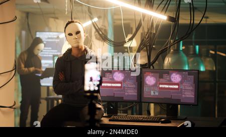 Weibliche Hacker mit Maske, die Live-Bedrohungsvideo sendet, um Lösegeld bittet, anstatt wichtige Informationen zu verbreiten. Gefährliche maskierte Frau droht, Daten preiszugeben, Cyberangriff. Stockfoto