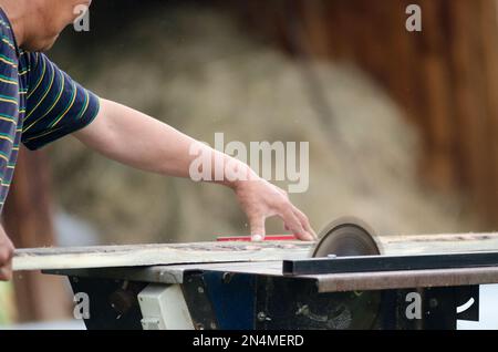 Ein Mann sägt ein Brett auf einer Kreissäge vor dem Hintergrund eines Schuppens mit Holz. Stockfoto