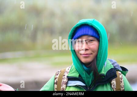 Yakut Asiatin Tourist mit Hut und Kapuze, lächelnd mit Brille vor dem Hintergrund von verschneitem Wetter und grünem Gras im Wald in der Nähe des Flusses im N Stockfoto