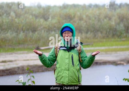 Yakut Asiatin Tourist in einer Jacke Hut und Kapuze und mit einem Rucksack schließt seine Augen, unter Hagel und Regen hält seine Hände gegen den Ba Stockfoto