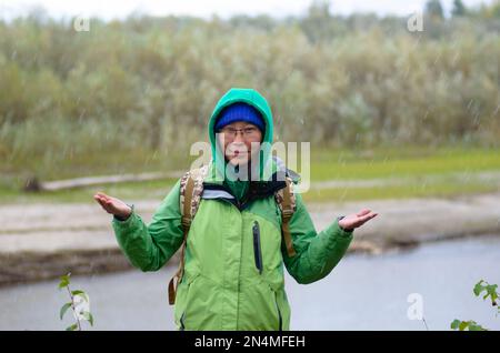 Yakut Asiatin Tourist in einer Jacke Hut und Kapuze und mit einem Rucksack lächelt, Hagel und Regen halten seine Hände vor dem Hintergrund des Wil Stockfoto