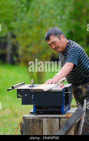 Yakut-Mann mit angespanntem Gesichtssägebrett auf einer Kreissäge vor einem grünen Garten im nördlichen Dorf. Stockfoto