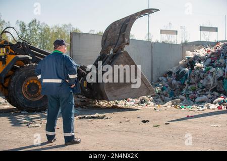 Ein Arbeiter im Recycling-Center, in dunkelblauer Arbeitskleidung, der sich einen unsortierten Müllhaufen ansieht, ist gerade in der Materialrückgewinnungsanlage angekommen, Rückansicht. Stockfoto