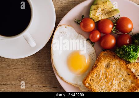 Schließen Sie das hausgemachte Frühstück mit einer Tasse schwarzem Kaffee, Spiegelei und Toast mit Tomaten und Avocado auf einem Teller Stockfoto