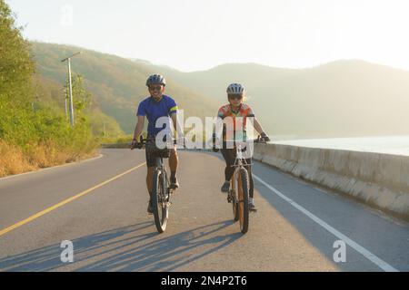 Asiatisches Fahrradpaar, das am Morgen gemeinsam um den See reitet, mit wunderschönem Blick auf die Berge im Hintergrund. Stockfoto