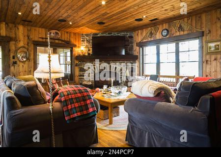 Wohnzimmer mit Holzdecke und -Wänden, grau-braunen Polstersofas, antikem Holztisch, Schaukelstuhl, Natursteinkamin. Stockfoto