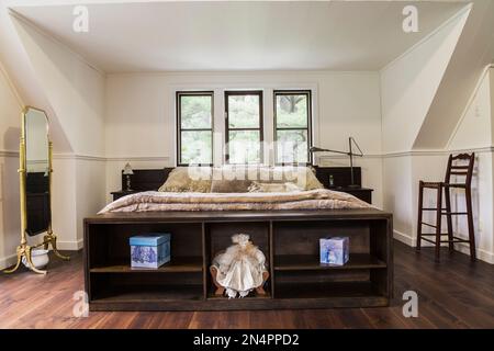 Holzrahmen Kingsize-Bett mit blumigem Bettüberwurf, Messingspiegel und antikem hölzernen Hochstuhl im Hauptschlafzimmer im New Hampton Stil Haus. Stockfoto