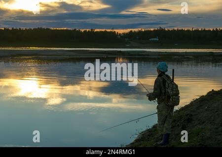 Ein weiblicher Fischer mit einem Rucksack, der eine Stange in den Beinen hält, wirft eine zweite Drehung vor dem Hintergrund eines hellen Sonnenuntergangs in der wilden Taiga von Stockfoto