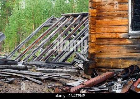 Eine Ecke eines abgebrannten Holzhauses mit einem Fenster und einem Gewächshausdach neben gelbem Holz und schwarz verkohlten Holzstämmen im Wald nach dem Feuer. Stockfoto