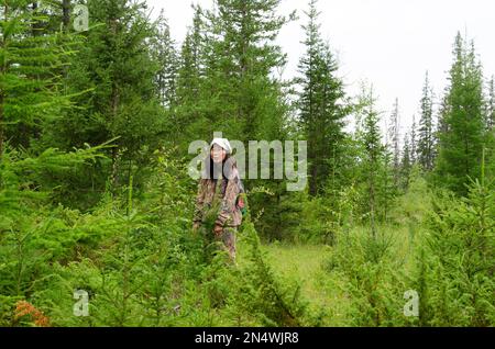 Yakut asiatische Touristin mit einem Rucksack in Schutzkleidung steht in den Büschen gedanklich im wilden Wald der Fichtentundra des Nordens. Stockfoto