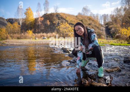 Yakut-Asiatin in Brille, lächelnd, sitzt am Wasser und berührt die Oberfläche. Stockfoto