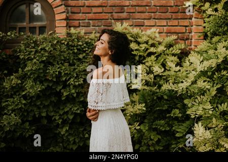 Hübsche junge Frau mit lockigem Haar steht in einem wunderschönen Garten, gekleidet in einem weißen Kleid. Sie sieht entspannt und zufrieden aus Stockfoto