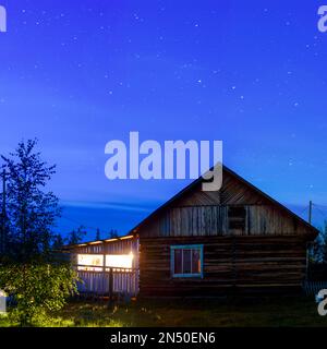 Das helle Licht der Lampe von der Veranda eines Privathauses beleuchtet die Birke und das Gras, die nachts unter dem Himmel stehen, mit Sternen im Himmel Stockfoto