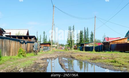 Heller Tag im Dorf ulus Suntar in Yakutia an einer Wohnstraße mit einer großen Pfütze und einem Auto in der Ferne zwischen den Holzhäusern. Stockfoto