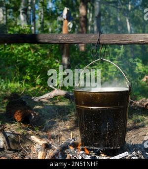 In einem Eimer, der an einem Stock über einem Feuer hängt, wird Suppe im wilden nördlichen Wald von Yakutia am Nachmittag inmitten der Vegetation gekocht. Stockfoto
