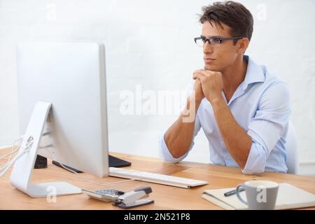 Analyse der Zahlen... Ein gutaussehender junger Geschäftsmann mit Brille, der aufmerksam auf seinen Computerbildschirm blickt. Stockfoto