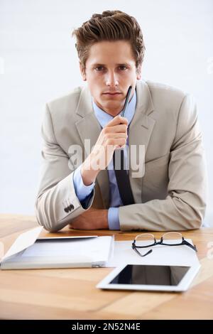 Intensiver Geschäftsmann. Porträt eines seriösen jungen Geschäftsmanns, der mit einem Tablet und Papierkram an seinem Schreibtisch sitzt. Stockfoto