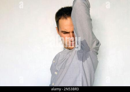 Der Mann schnüffelt verschwitzte Achseln auf blauem Hintergrund. Unangenehmer Schweißgeruch. Unzufriedener Gesichtsausdruck. Nasser Fleck auf meinem blauen Hemd durch Schweiß. Stockfoto