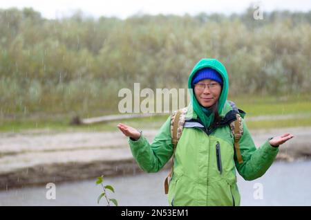 Yakut Asiatin Tourist in einer Jacke mit Hut und Kapuze und mit einem Rucksack lächelt, Hagel und Regen halten seine Hände vor dem Hintergrund des riv Stockfoto