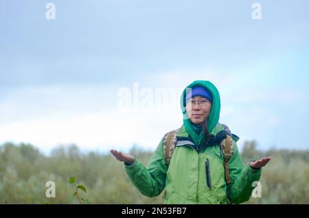 Yakut Asiatin Tourist in einer Jacke Hut, Kapuze und Brille, mit einem Rucksack lächelt, Hagel und Regen hält seine Hände vor den Hintergrund von t Stockfoto