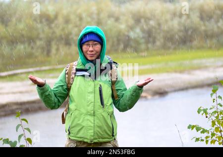 Yakut Asiatin Tourist in einer Jacke mit Hut und Kapuze und mit einem Rucksack lächelt, Hagel und Regen halten seine Hände vor dem Hintergrund des riv Stockfoto