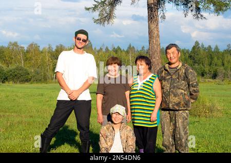 Ein großer russischer Mann in Stiefeln steht neben einem kurzen asiatischen Yakuts und ein Mädchen, das auf dem Gras sitzt und auf einem Feld im wilden Wald des Nordens für ein Foto posiert Stockfoto