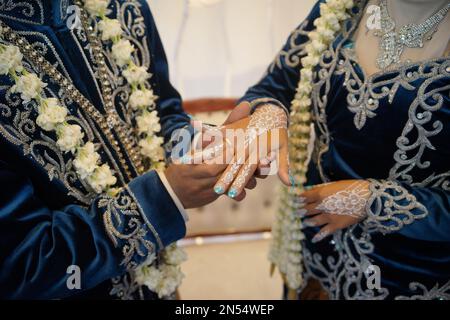 Der Bräutigam legt den Hochzeitsring auf die Hand der Braut und trägt in einer traditionellen Hochzeitszeremonie ein Hochzeitskleid mit Jasmine- und Magnolienblumen Stockfoto