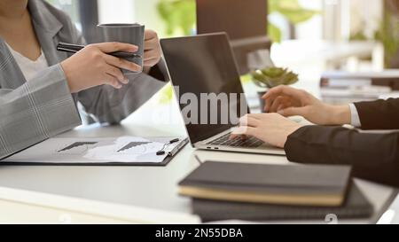 Zugeschnittenes Bild von zwei Geschäftsfrauen, die in einem gemeinsamen Arbeitsraum zusammenarbeiten, ein Notebook verwenden, diskutieren, Brainstorming durchführen und während der Arbeit Kaffee trinken. Stockfoto