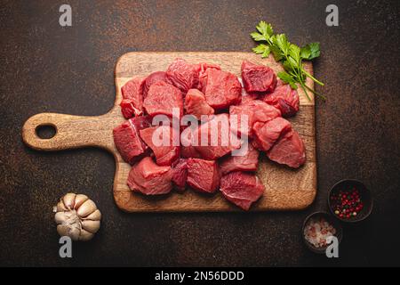 Rohes Rindfleisch in Würfel gehackt mit einem Haufen frischer Petersilie, Knoblauch, Salz und Pfeffer auf einem Holzbrett zum Kochen von Eintopf oder Fleischgericht auf Braun Stockfoto