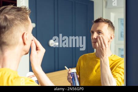 Ein erwachsener Mann hält die Tonic-Flasche im Gesicht und trägt das Produkt mit einem Wattepad auf. Männliches Porträt im Spiegelwischgesicht. Hautpflege Morgenroutine, vorsichtige Schönheit Stockfoto