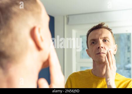 Ein erwachsener Mann trägt Gesichtscreme mit Wattepad auf und massiert dann mit den Händen. Männliches Porträt im Spiegel. Hautpflege Morgenroutine, sorgfältige Schönheitsbehandlung Stockfoto