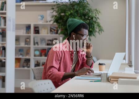 Fokussierter junger afrikanischer Lehrer, der sich mit einem Laptop umsieht, um sich auf den Online-Kurs in der Bibliothek vorzubereiten Stockfoto
