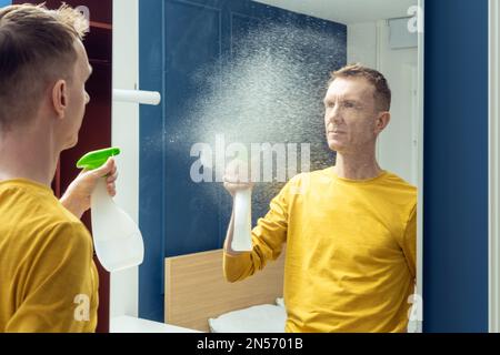 Ein Mann mittleren Alters hält eine Flasche Reinigungsmittel und spritzt Spritzer auf den Spiegel der Schiebetür. Spiegel mit Haushaltsreiniger waschen und polieren. Alles Klar Stockfoto