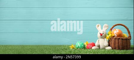 Farbenfrohe Ostereier, Korb und Hasenspielzeug auf grünem Gras auf hellblauem Holzhintergrund, Platz für Text. Horizontales Bannerdesign Stockfoto