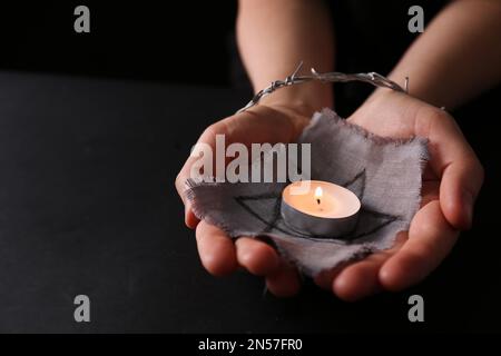 Frau gefesselt mit Stacheldraht, mit David-Stern und brennender Kerze auf schwarzem Hintergrund, Nahaufnahme. Holocaust-Gedächtnisfeiertag Stockfoto