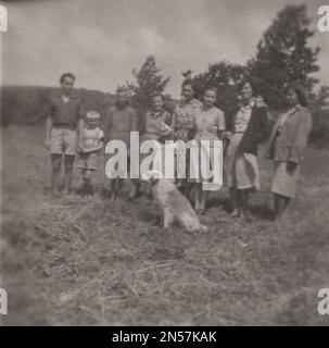 Vintage-Foto, auf dem die Familie mit ihnen das Hündchen liebt / Vintage-Erinnerung / gute alte Zeiten: Die ganze Familie steht in einer Reihe für die Kamera und natürlich muss in der Mitte der liebende Hund der Familie im 1930er sein Stockfoto