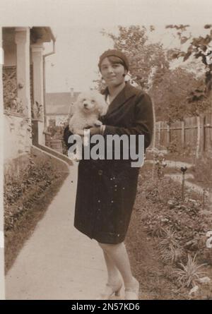 Vintage-Foto von Lady und ihrem Hund / Hündchen in der Hand / Hunde umarmen / Hunde umarmen / Hund unter den Armen halten. Junge Dame posiert für die Kamera mit ihrem süßen weißen Spielzeughund. Possibli der Welpe ist ein Spielzeugpudel oder Coton de Tulear, Bolognese oder sogar Malteser Stockfoto