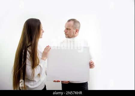 Ein erwachsener Mann und eine junge Frau, die ein Bild in den Händen halten, Rahmen für Werbetexte, die aufrichtiges Lachen lächeln, weiße Kleidung, weißer Hintergrund, leerer Rahmen, Papier, Pappe, Posterbanner Stockfoto