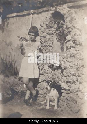 Vintage-Foto von Lady und ihrem Hund/jungen Dame mit ihrem Jack Russell Terrier vor einem Jugendstilbrunnen/Jugendstil-Lady-Gesichtsskulptur in einem versteckten Garten, einem geheimen Garten / um 1930er Uhr. Stockfoto