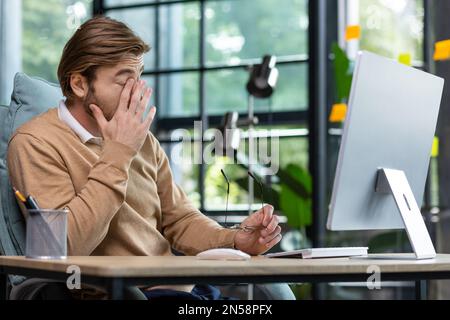 Müde Büroangestellte reiben die Augen nach langer Arbeit am Computer im Büro, junger blonder Geschäftsmann, der in lässiger Kleidung am Arbeitsplatz überarbeitet wurde. Stockfoto