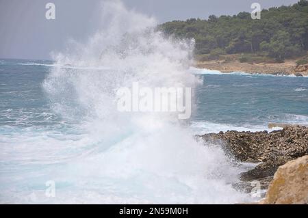 Wellenspritzer im Meer gegen das Strandriff, Wellenspritzer. Mächtige Wellen an einem felsigen Strand. Wellen, die in die Felsen stürzen, Insel Losinj, Kroatien Stockfoto